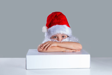 Little Santa Claus put his head on his folded arms on the white box. He is sitting at a white table. Sad looking at the camera. Gray background. Close-up.