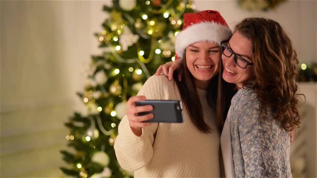 Two Women Taking a Selfie Having Fun at Home. Brunette Sisters Using Smartphone and Photographing Themselves on Christmas Tree Background. One Girl Wearing Glasses, Another - Santa Hat.