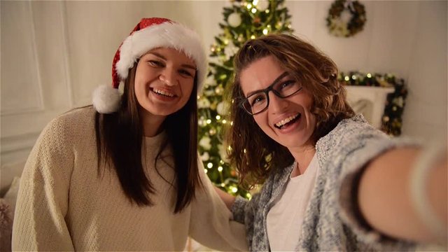 Two Smiling Attractive Women Making Selfie on Christmas Tree Background. One Girl with Curly Hair Wearing Glasses, Another - Santa Hat.