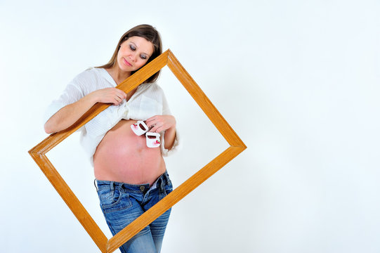 Pregnant girl holding picture frame from picture and children's shoes near naked belly on white background. Portrait. Landscape orientation
