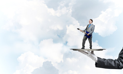 Businessman on metal tray playing electric guitar against blue sky background