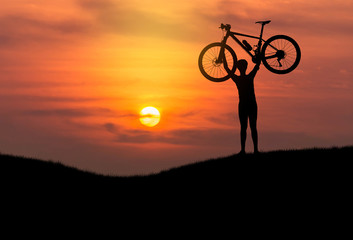 Silhouette the man stand in action lifting mountain bike bicycle above his head on the meadow with sunset