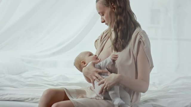 Young beautiful mother sitting on bed and breastfeeding and talking to her fussy baby boy 
