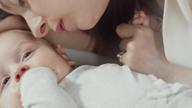 Handheld shot of little baby boy lying on bed sucking on hand as his mother gently kissing him and talking 