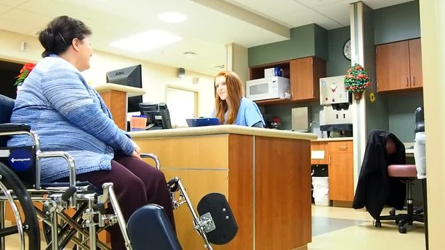 Female nurse taking patient information while patient sits in wheelchair at nurses station
