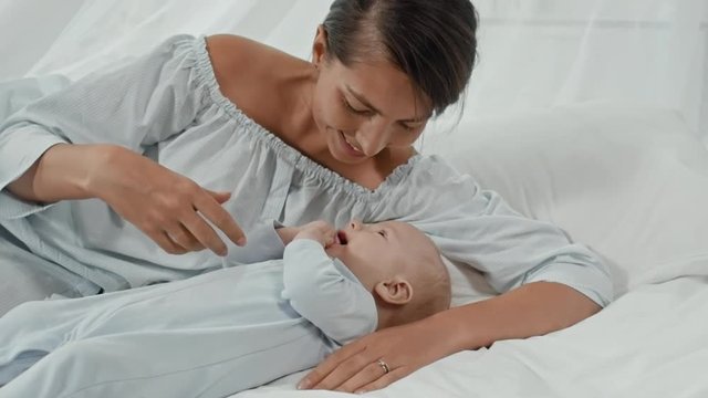 Young mother lying in bed and cuddling infant son, then caressing him and kissing his nose