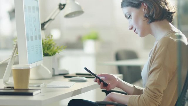 Beautiful Brunette Uses Smartphone While Sitting at Her Light and Modern Office. Shot on RED Cinema Camera in 4K (UHD).