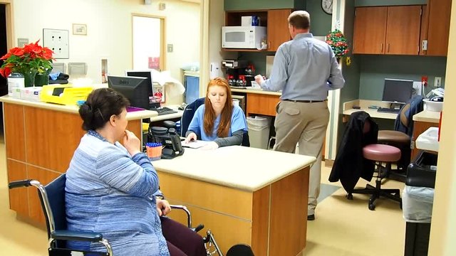 Doctor talking with nurse and patient at nurses station while patient sits in wheelchair.
