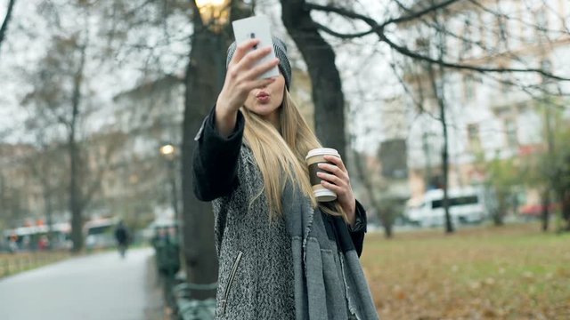 Happy girl in warm clothes doing selfies on smartphone in the autumnal park, steadycam shot
