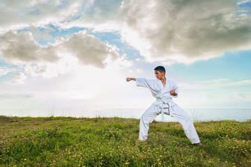 Fototapeta na wymiar Boy in white kimono during training karate exercises in summer outdoors with sky and sea background.