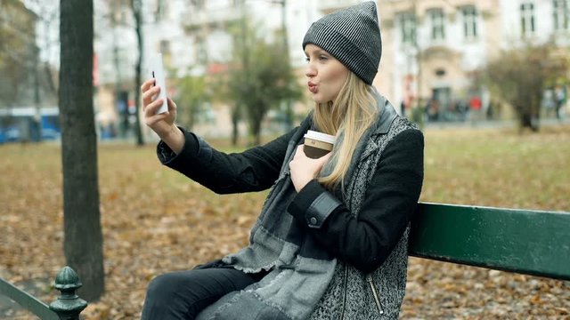 Girl sitting in the autumnal park and doing selfies on smartphone, steadycam shot
