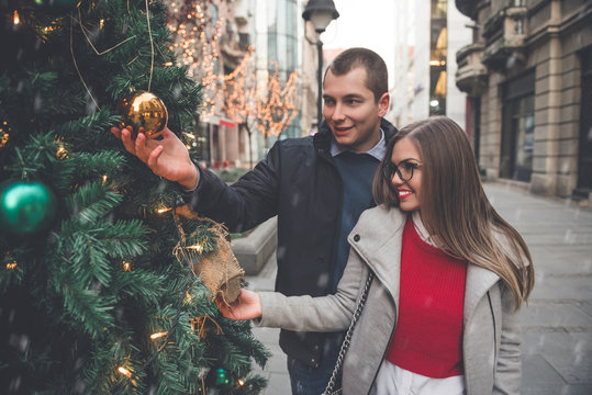 Beautiful young couple enjoying in cold winter evening. They standing on the street and looking ornaments on Christmas tree. Colorful street lighting in background. New Year theme.