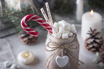 Cocoa with marshmallow, candy cane and straws in the glass jar decorated with little white heart on the table with winter decor for holidays. Close up