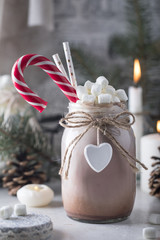 Cocoa with marshmallow, candy cane and straws in the glass jar decorated with little white heart on the table with winter decor for holidays. Close up