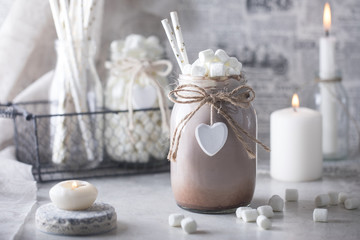 Fototapeta na wymiar Cocoa with marshmallow and straws in the glass jar decorated with little white heart on the table with candles for the winter holidays