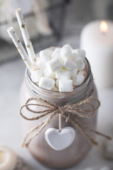 Cocoa with marshmallow and straws in the glass jar decorated with little white heart on the table with candles for the winter holidays