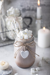 Cocoa with marshmallow and straws in the glass jar decorated with little white heart on the table with candles for the winter holidays