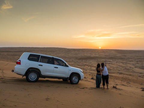 Oman, Al Raka, two young women standing besides off-road vehicle on a desert dune watching sunset