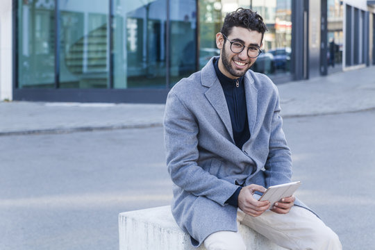 Portrait of smiling young man with mini tablet sitting on bollard