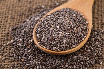 Wooden bowl full with chia seeds and a spoon next to it on sack cloth background.