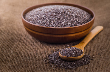 Wooden bowl full with chia seeds and a spoon next to it on sack cloth background.