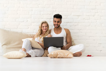 Young Couple Sit On Pillows Floor, Hispanic Man And Woman Using Laptop Computer Lovers Bedroom Over White Brick Wall