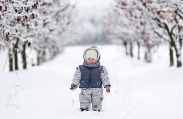 Fototapeta na wymiar Little kid in snowsuit, hat and scarf, walking through a snowy path