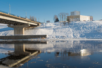City winter landscape, ice floes floating on the river, the steep bank of snow, bridge, buildings, trees and sky reflected in the river