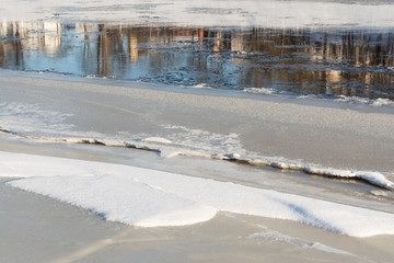 bank of the river is covered with ice, gray and white snow, but in the middle of the river continues to flow, shows the reflection of the city
