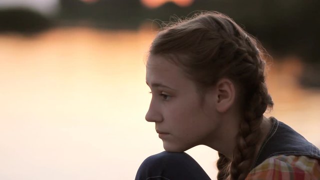 Sad teen girl beach. Side view of thoughtful pretty girl resting her chin on her knee while relaxing by calm river at sunset.