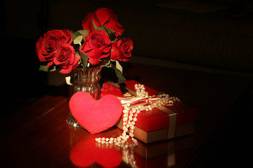 Background for St. Valentine’s’ Day. Box of delicious candy decorated in red and gold colors with pearl necklace next to the vase with red roses and red heart on the foreground on the wooden table