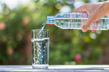 Man's hand holding plastic bottle water and pouring water into glass on wooden table on blurred...