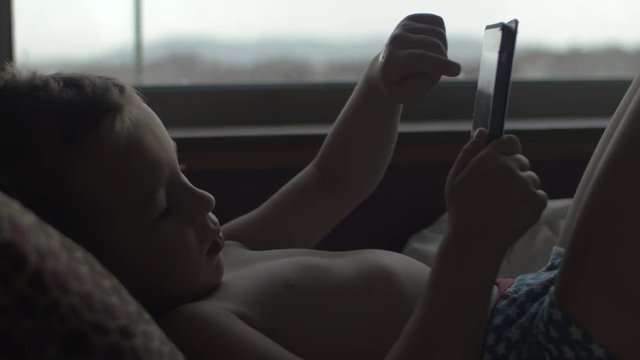 Boy in pants lying in bed by the window and using tablet computer. Resting with touch pad
