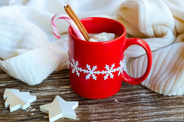 Red mug of hot chocolate with cinnamon stick  and candy cane, white cozy knitted sweater on the back. Delicious cold weather beverage. Winter times or Christmas morning concept.
