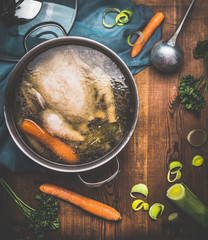 Chicken soup cooking, pot with chicken broth and  ladle on dark rustic wooden background with...