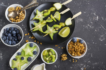 Healthy fresh breakfast. Granola, quinoa bowl, various nuts and fruit skewers with green fruits on a dark background. Top view. Color year. Greenery. 