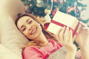 Beautiful happy young woman lying on the sofa holding a Christmas present