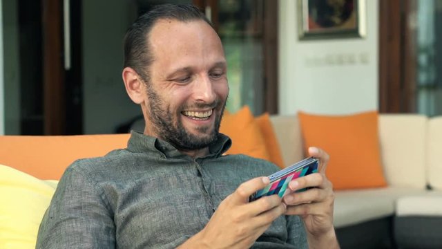 Young, happy man playing game on smartphone lying on lounger on patio
