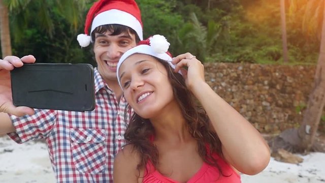 Happy couple on Christmas travel holidays taking selfie picture on a tropical beach with smartphone wearing santa hat during sunset in slow motion. 1920x1080