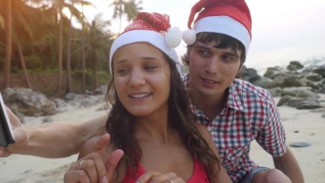 Happy beautiful couple on Christmas travel holidays taking selfie picture on a tropical beach with smartphone wearing santa hat during sunset in slow motion. 1920x1080