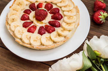 Tart with strawberries, bananas and whipped cream on a background of flowers. Top view. Close-up