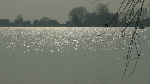 A great lake at the end of a sunny day. Light glimmers delicately on the surface of the water. In the background a few houses and trees.