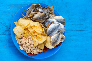 Close up of the mix beer snacks on wooden background. Potato chips, roasted and salted peanuts, dried fish in blue plate on the blue wood background.Top view/The beer snacks