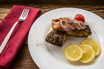Fried fish fillet with lemon and tomatoes on a white plate. Wooden background. Top view. Close-up