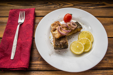 Fried fish fillet with lemon and tomatoes on a white plate. Wooden background. Top view. Close-up