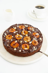 Closeup of a whole homemade Sticky Chocolate Plum Cake on a porcelain plate with a cake server on white background. In the background a cup of coffee, plate and milk jug. Shallow depth of field. 