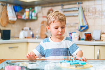 Little blond kid plays with molds for making ginger biscuits or cookies, sitting at the kitchen table with raw dough sheet and wheat flour. Duck pursed lips.