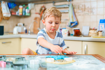 Fototapeta premium Little blond kid plays with molds for making ginger biscuits or cookies, sitting at the kitchen table with raw dough sheet and wheat flour. Protruding lower lip.