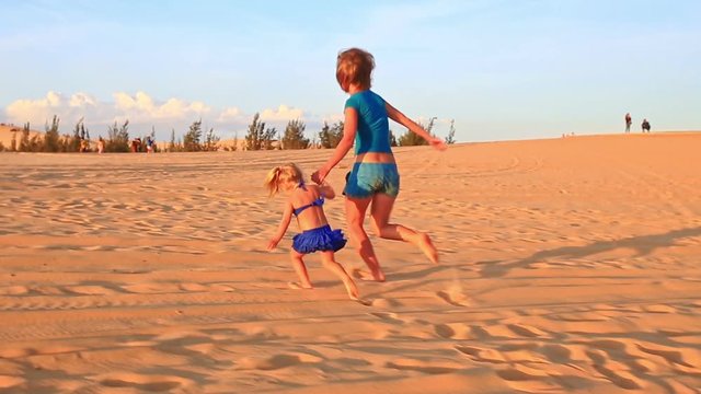 backside caucasian mother and small blond girl with pigtail join hands run barefooted on sand dunes at sunset