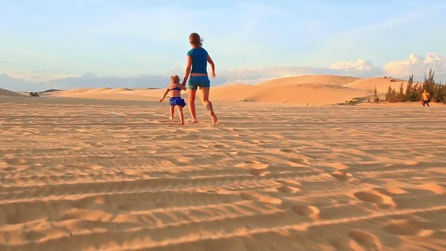 backside caucasian mother small blond girl join hands walk barefooted on sand dunes jeep crosses way at sunset
